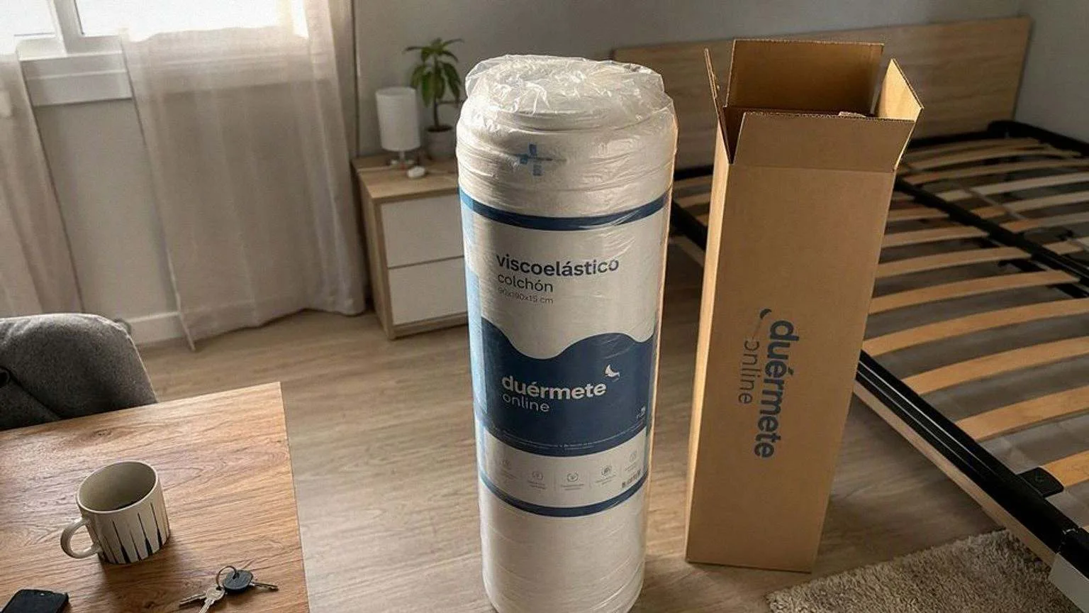 Colchón viscoelástico enrollado y precintado al vacío, de pie junto a su caja de cartón abierta en un dormitorio a medio amueblar